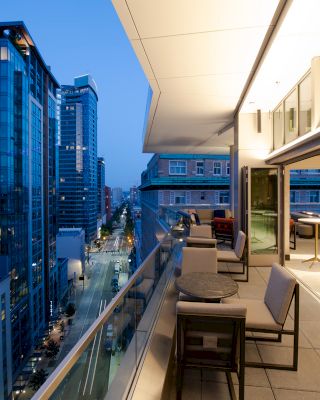 The image shows a modern cityscape at dusk with high-rise buildings. In the foreground, there's a balcony with seating and tables.