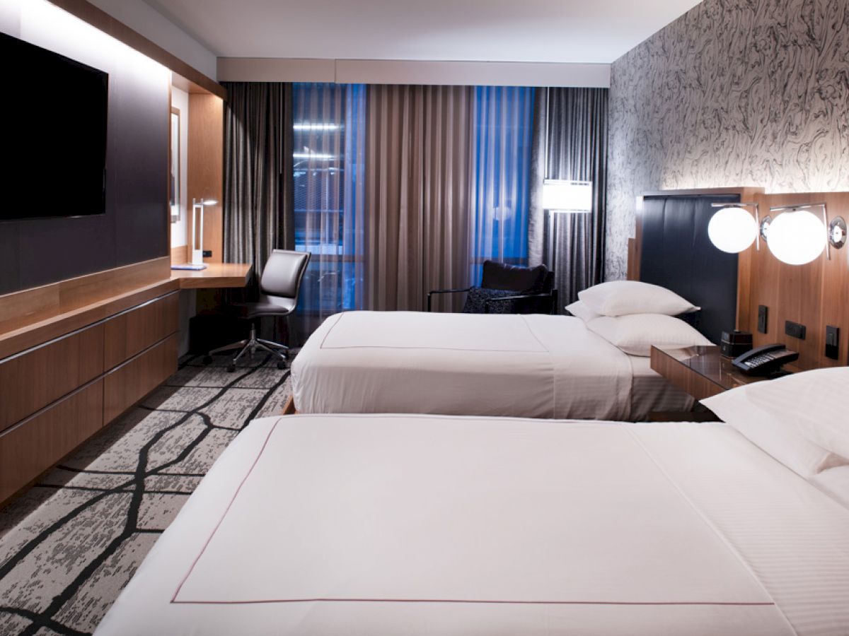 A modern hotel room features two beds, a wall-mounted TV, a desk with a chair, and a lamp. The decor is neutral with a patterned wall.