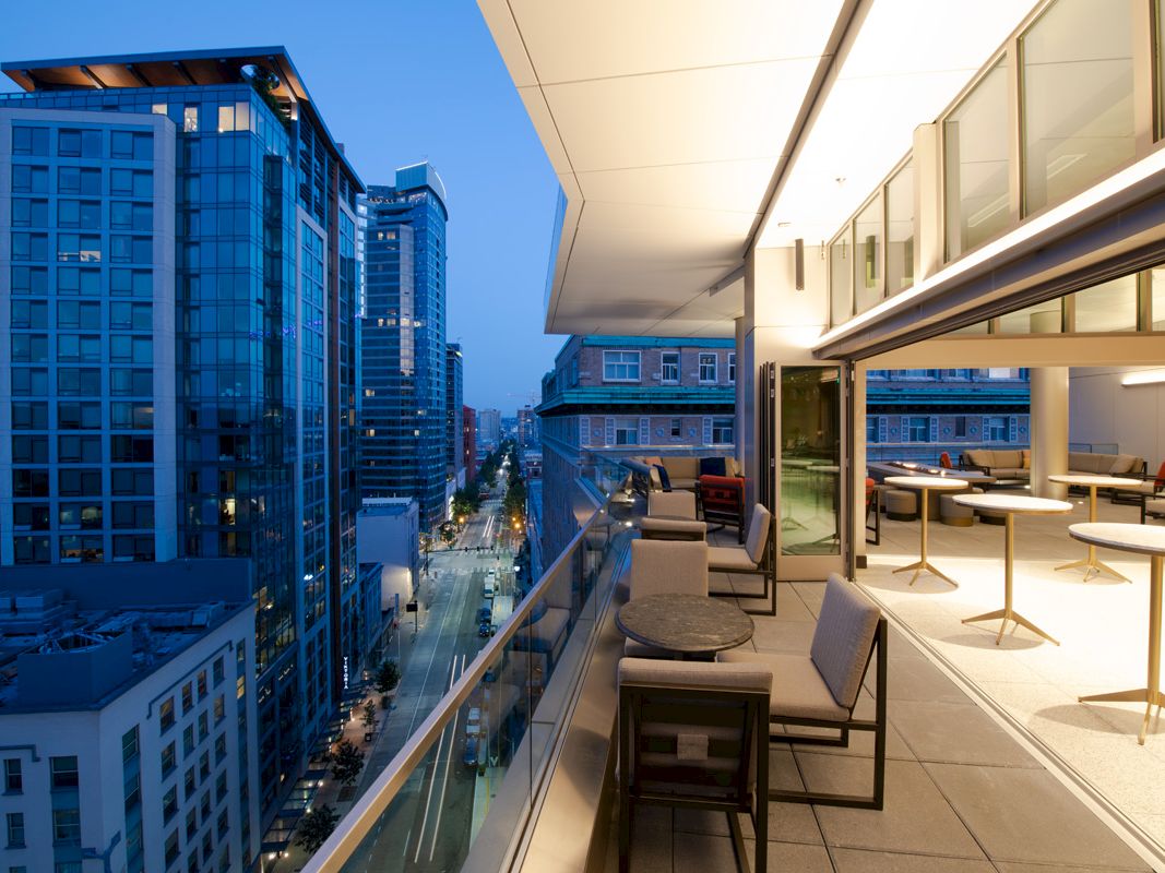 A modern cityscape at dusk with tall buildings, viewed from a spacious, furnished balcony with seating and tables, adjacent to large windows.