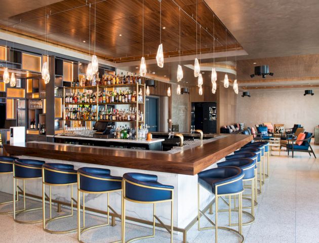 The image shows a modern bar with a stylish interior, featuring a wooden counter, cushioned bar stools, hanging lights, and a well-stocked liquor shelf.