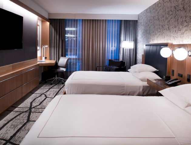 A modern hotel room with two beds, a TV, a desk, and large windows covered by curtains. The walls have decorative patterns.