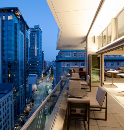 A modern rooftop balcony at dusk with cityscape views, featuring tables, chairs, and large windows of nearby buildings illuminated by lights.