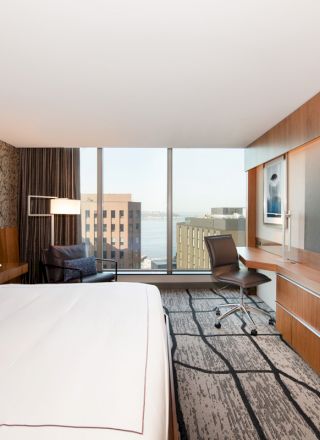 A modern hotel room with a large bed, a wall-mounted TV, a work desk with a chair, and a window offering a cityscape view is shown.