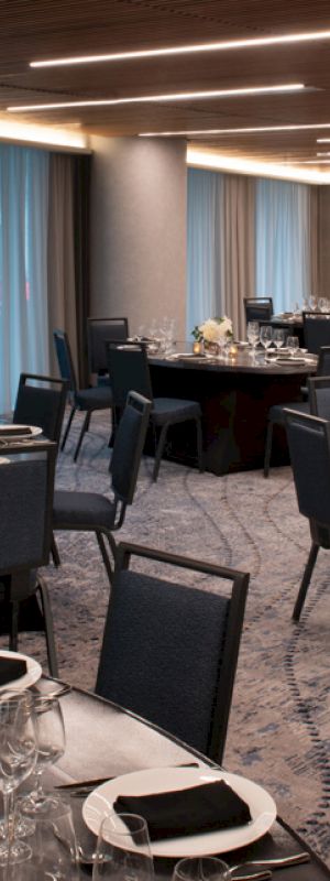 The image shows a modern, elegantly set dining room with round tables, black chairs, and floral centerpieces, all under a ceiling with linear lighting.