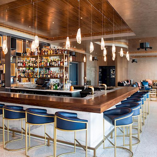 This image shows a modern bar with a U-shaped counter, blue bar stools, a variety of liquor bottles on shelves, and hanging pendant lights.