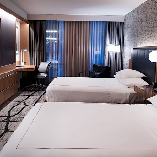 The image shows a modern hotel room with two beds, a desk, a TV, and lamps. The decor is contemporary, featuring neutral colors and patterned wallpaper.