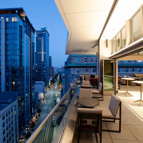 A modern balcony features seating areas overlooking a cityscape with tall buildings at dusk, creating a stylish urban ambiance.