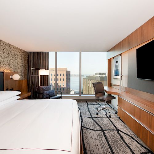 A modern hotel room features a large bed, flat-screen TV, desk, and chair, with floor-to-ceiling windows offering a city and partial waterfront view.