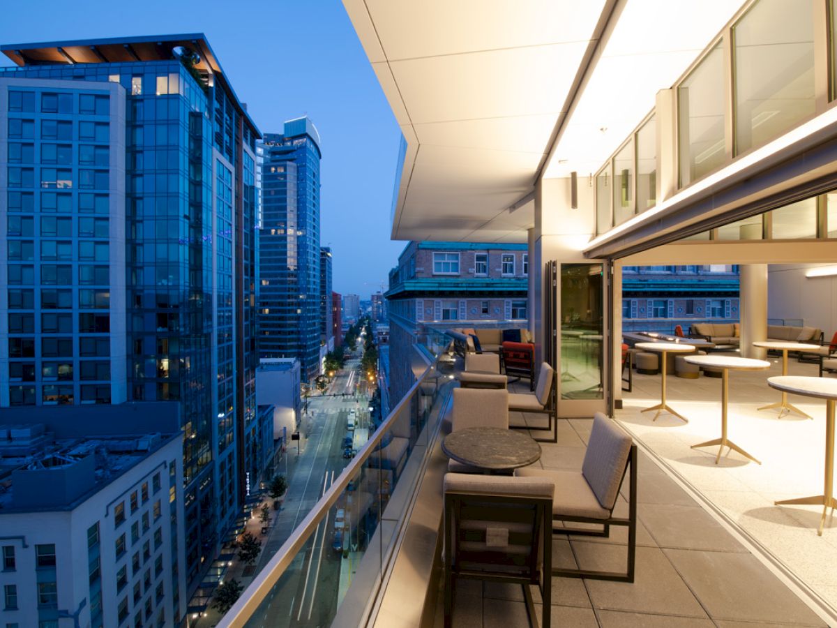 A modern outdoor balcony with tables and chairs overlooks a city street lined with tall buildings, illuminated by city lights at dusk.