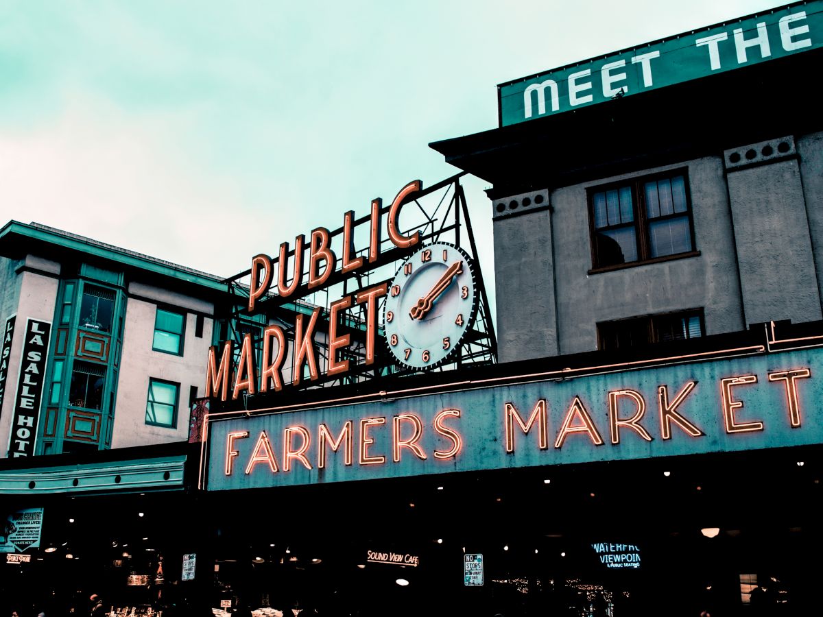 The image features a neon sign reading "PUBLIC MARKET" and "FARMERS MARKET" with a clock above, in front of buildings in an urban setting.