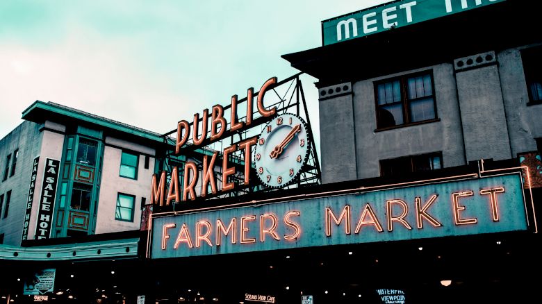 The image features a neon sign reading "PUBLIC MARKET" and "FARMERS MARKET" with a clock above, in front of buildings in an urban setting.