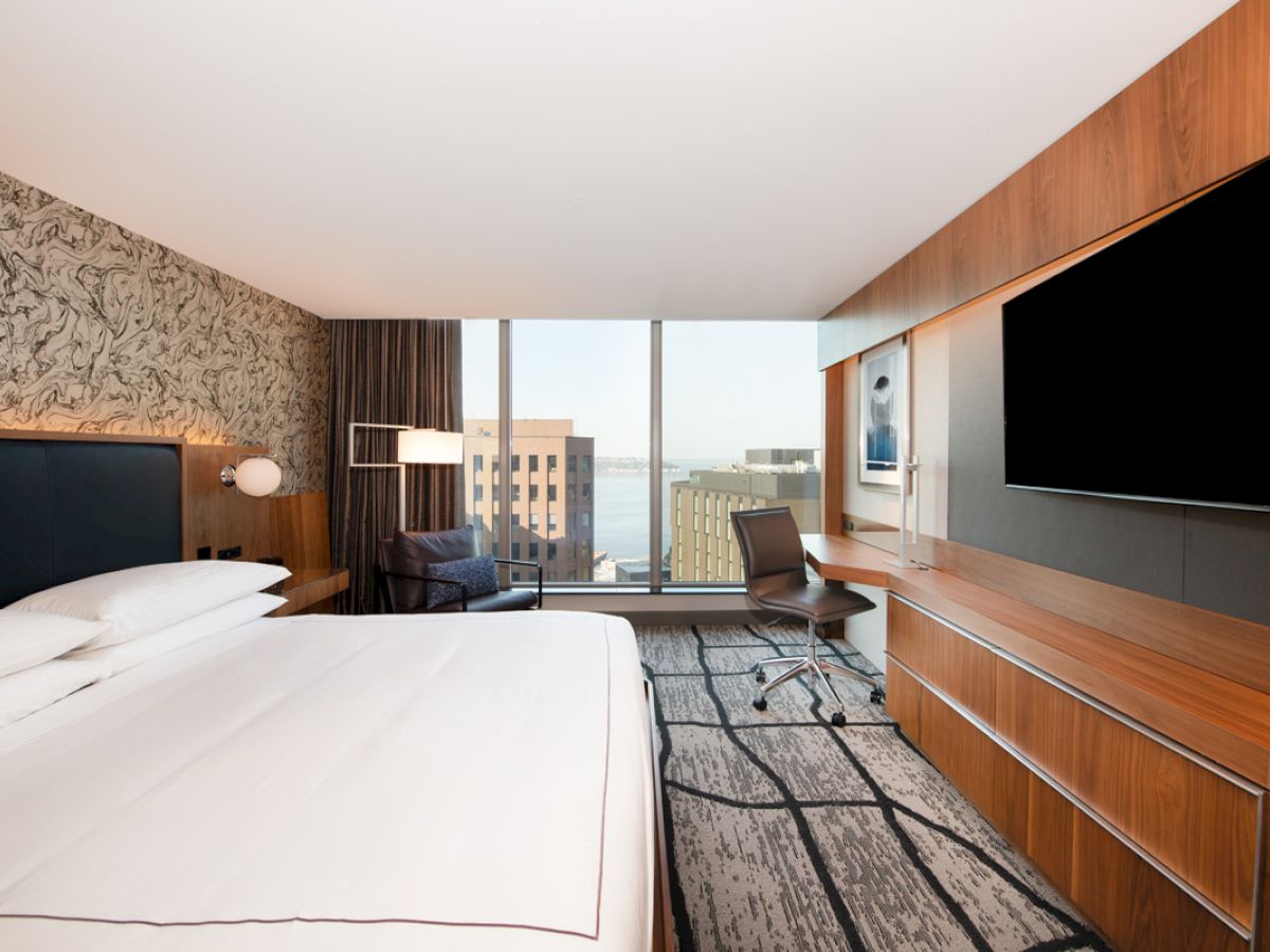The image shows a modern hotel room with a large bed, desk, chair, and flat-screen TV. There's a window with a cityscape view in the background.