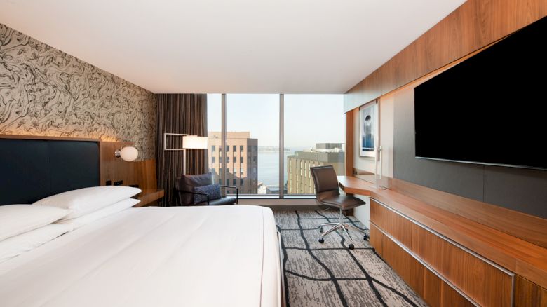 The image shows a modern hotel room with a large bed, desk, chair, and flat-screen TV. There's a window with a cityscape view in the background.