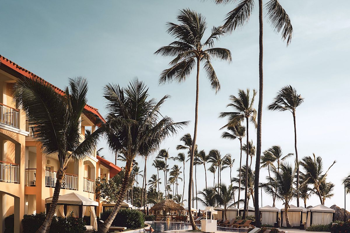 The image shows a serene tropical resort with a swimming pool, lounge chairs, tall palm trees, and a multi-story building with balconies, under a blue sky.