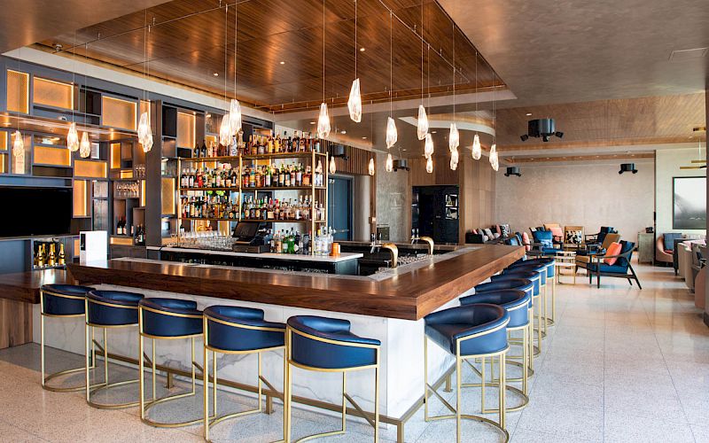 This image shows a modern, stylish bar with a wooden counter, blue upholstered stools, overhead pendant lights, and shelves filled with liquor bottles.