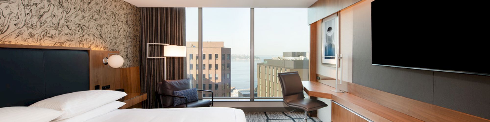 A modern hotel room with a large bed, desk, chair, wall-mounted TV, and floor-to-ceiling windows showing a cityscape view.