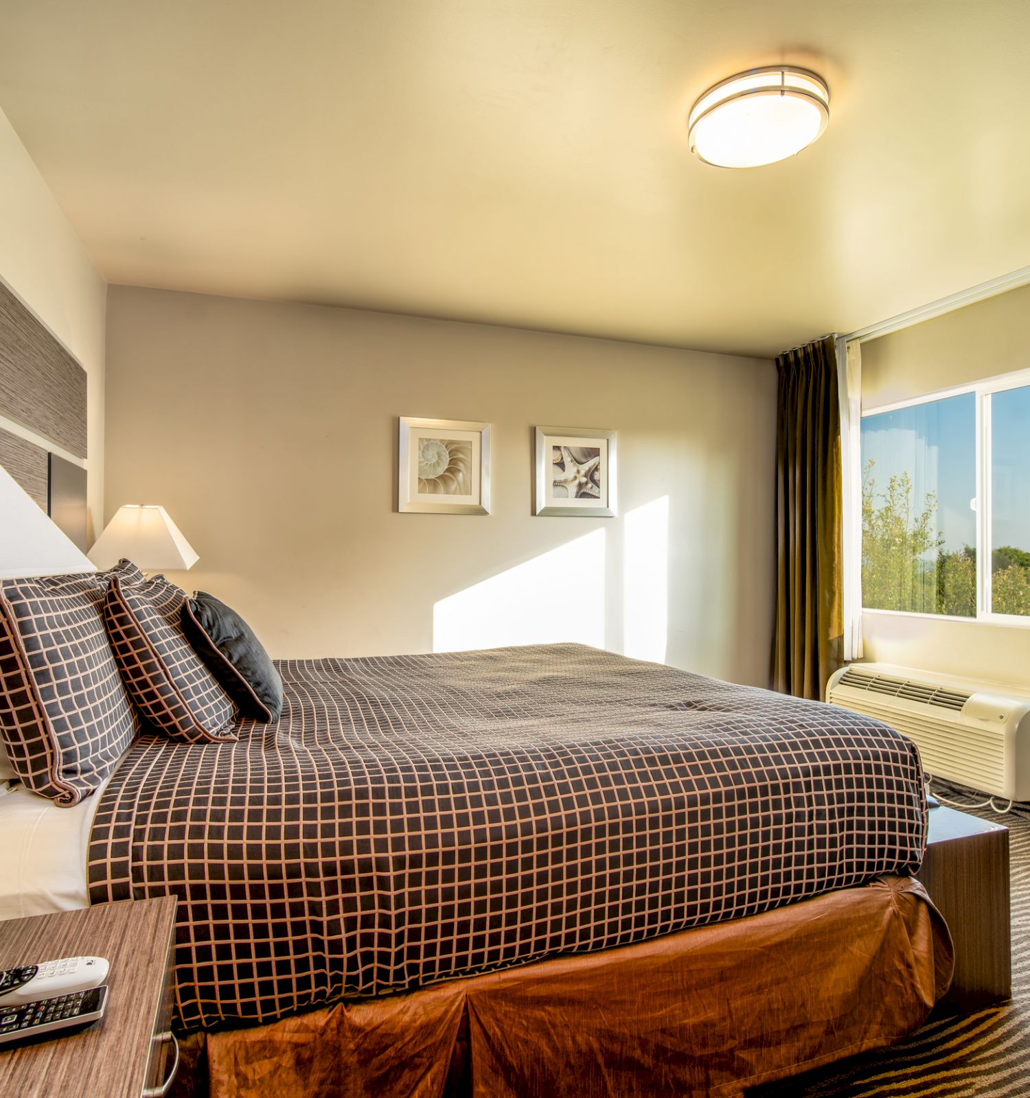 A modern hotel room features a large bed with checkered bedding, bedside lamps, framed art, and a window with a view, all in a cozy, well-lit space.