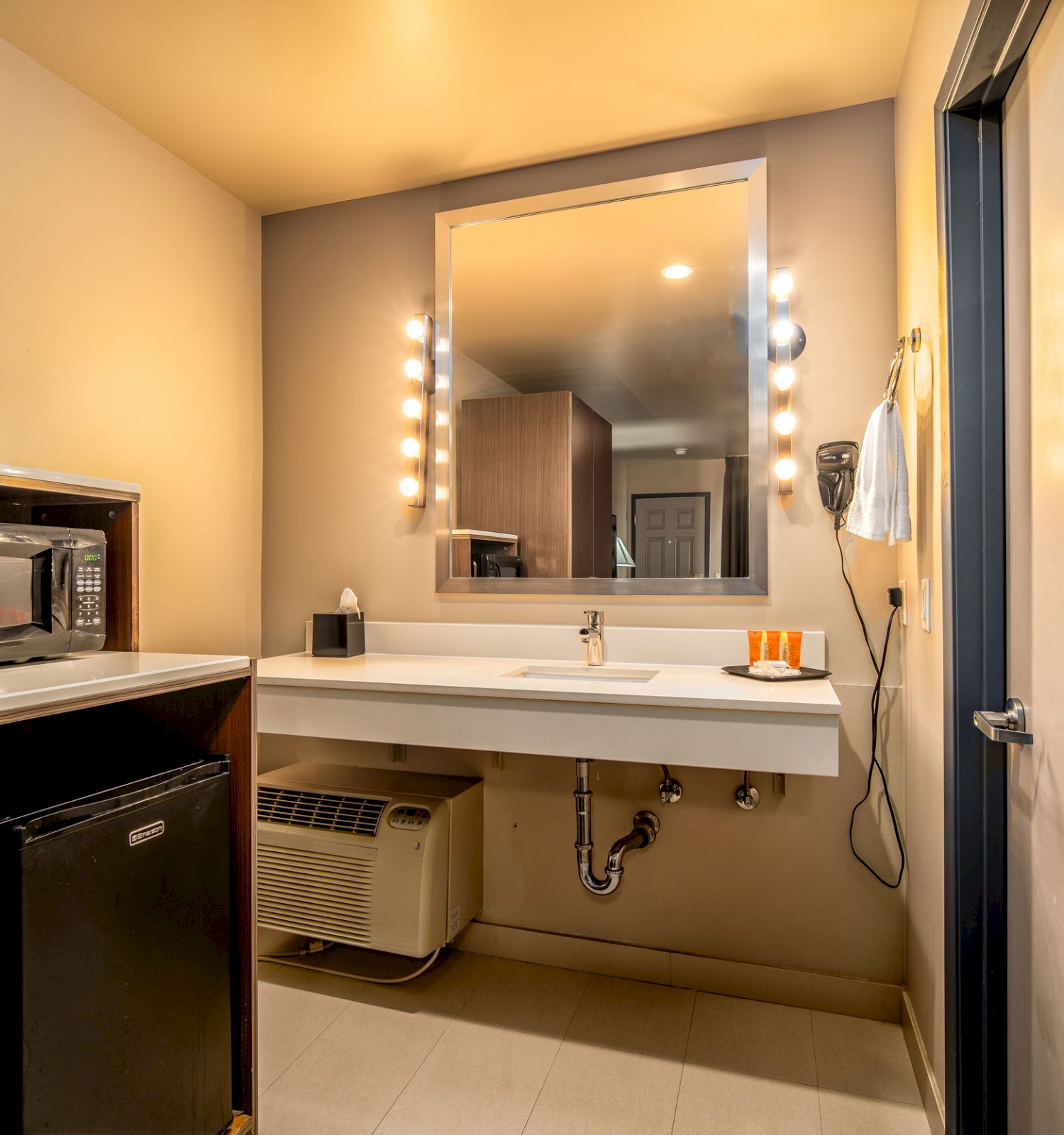 A modern bathroom with a large mirror surrounded by lights, sink, hairdryer, and toiletries on the countertop. A fridge and microwave are on the left.