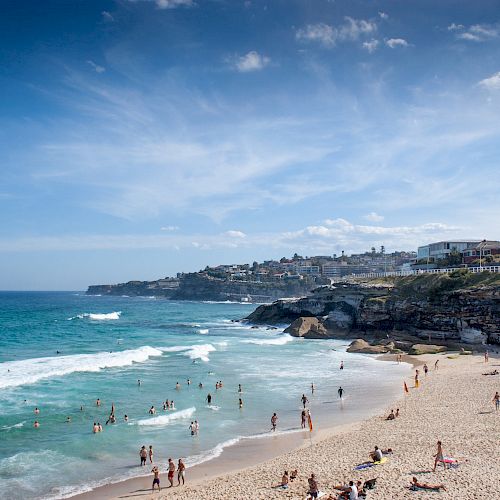 The image shows a scenic beach with people swimming, sunbathing, and enjoying the sunny day. There are buildings and cliffs in the background.