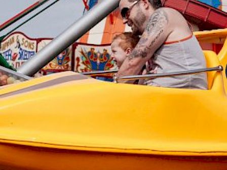 A man with tattoos and sunglasses enjoys a carnival ride with a young child in a bright yellow attraction car, amidst colorful fairground surroundings.