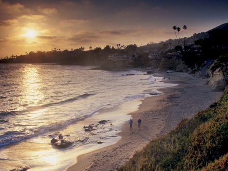 The image shows a serene beach at sunset with waves gently crashing, people walking along the shore, and houses and palm trees in the background.