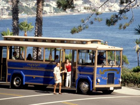 A vintage-style bus by a coastal area with people disembarking, surrounded by palm trees and ocean in the background.