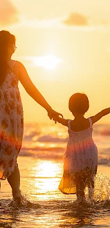 A woman and child holding hands, walking on the beach at sunset, with the sunlight creating a warm, golden glow.