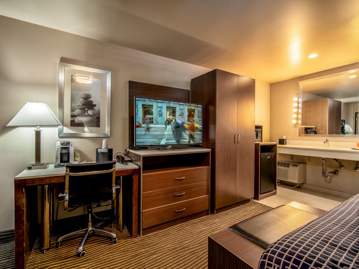 A hotel room features a TV, desk with chair, lamp, and framed picture. There's a wardrobe, cabinet, small fridge, bed, and adjacent bathroom area.